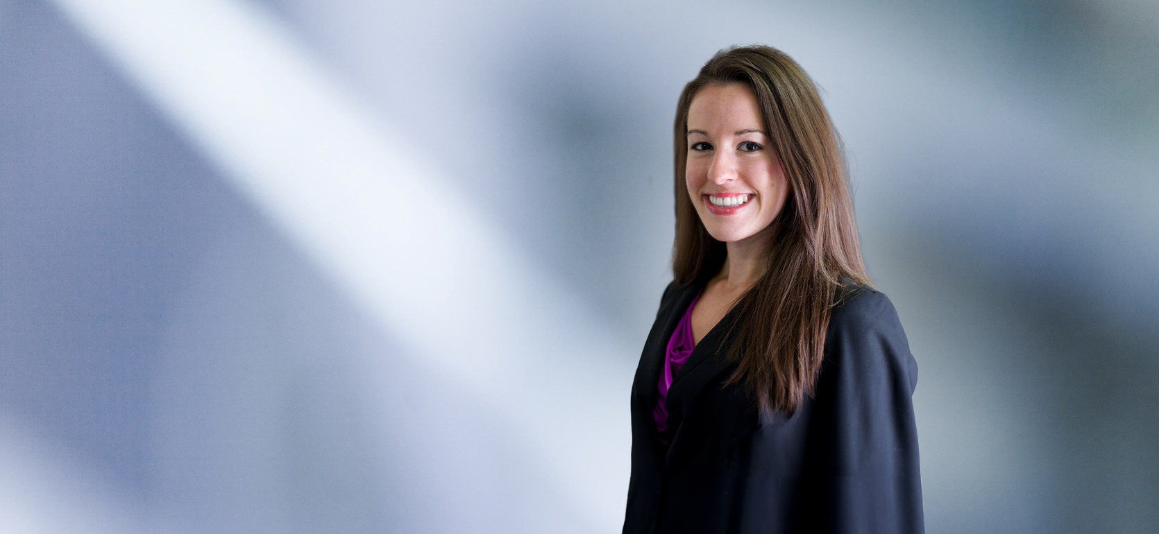 A woman, Julie Hamlet, in a black blazer and purple top is smiling at the camera, standing against a blurred blue and white background.