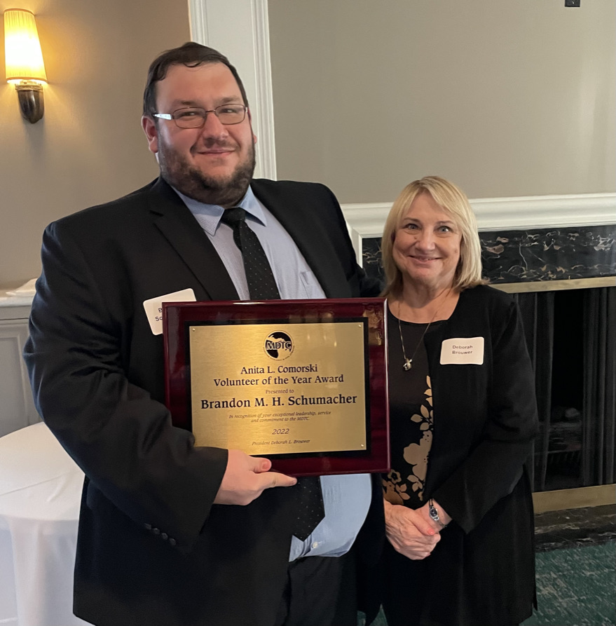 Brandon with Volunteer Award Brandon Schumacher in a suit being presented the Volunteer Award by a women in a black dress with blond hair