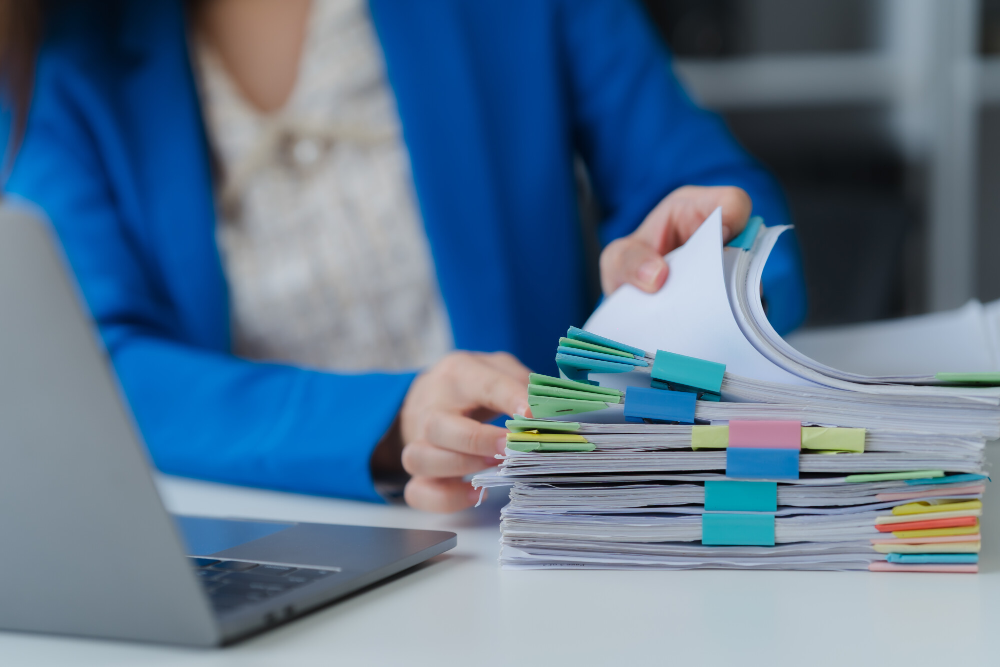 Workplace Documentation: A close-up shot of an office worker in a tailored blue blazer, meticulously reviewing a stack of documents. 