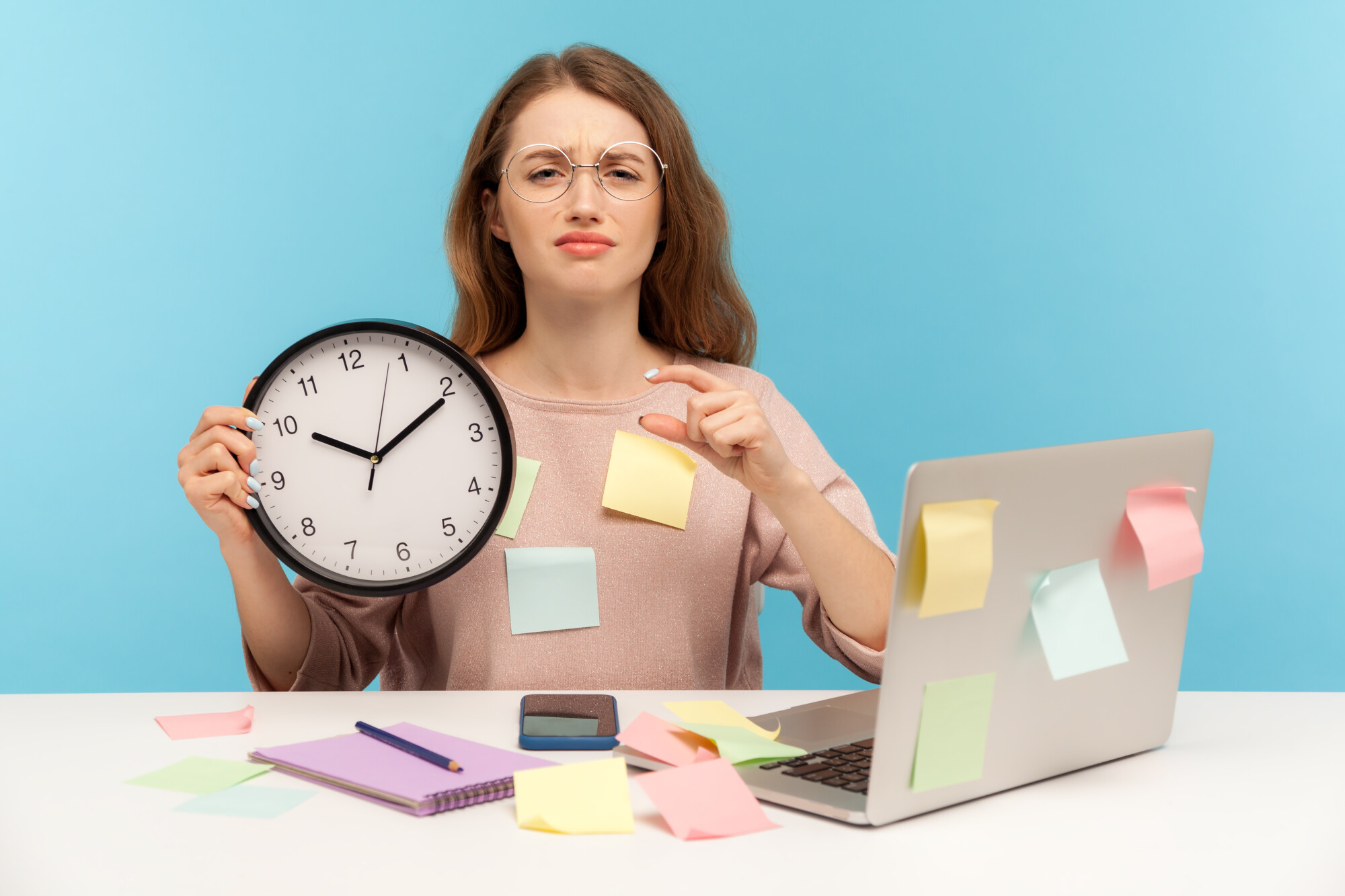 Exhausted fatigued upset woman employee sitting at workplace office, all covered with sticky notes, holding big clock and showing a little bit gesture, begging more time. 