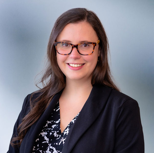 A brown-haired woman, Amy Marinkovski, wearing turtle frame glasses, black sport coat and black and white zigzag pattern shirt, smiles at camera in front of blue gray background