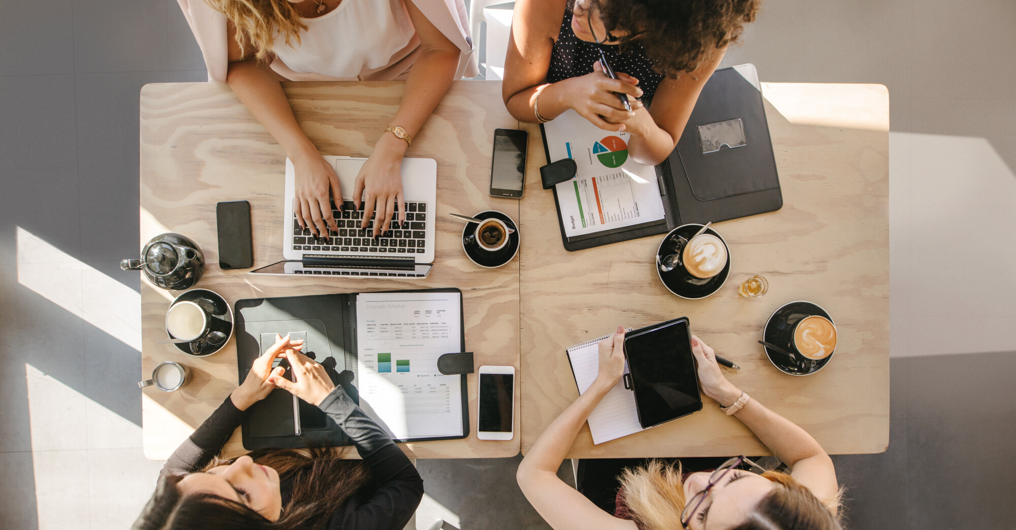 Four businesswomen sit around table with cups of coffee and laptops and documents
