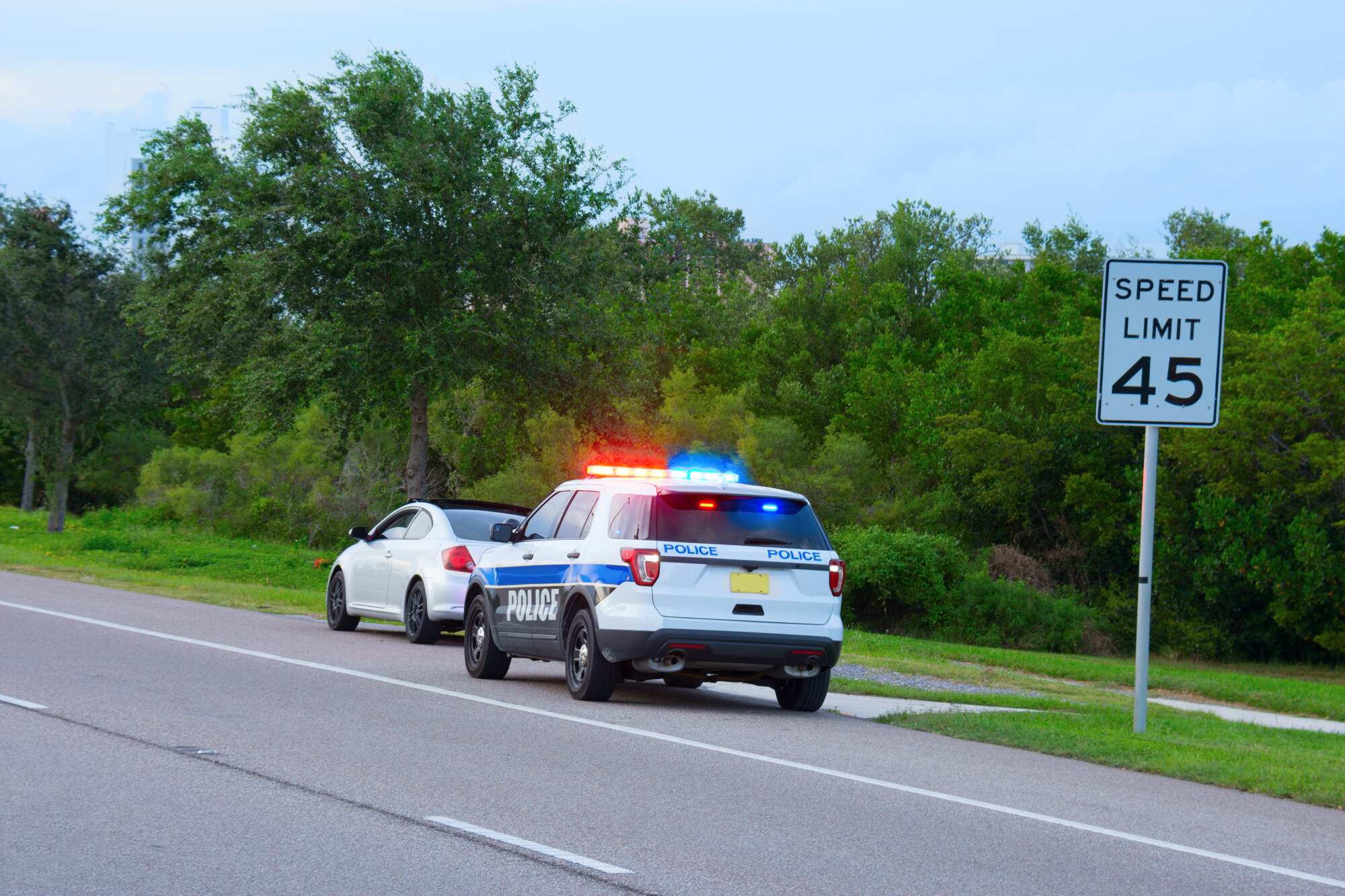 Police truck SUV vehicle with flashing red and blue lights has pulled over a sports car for speeding and they happen to be on the side of the road by a speed limit sign.