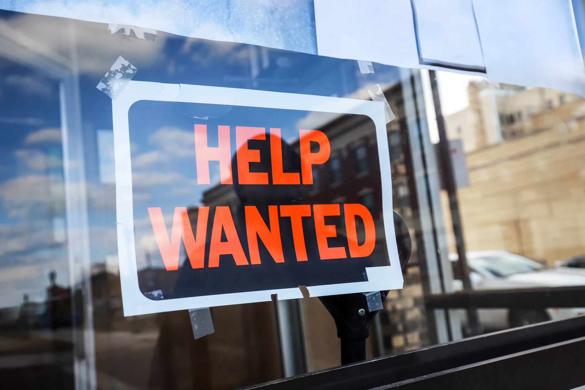 Black and White Sign with Orange Letters in the window of a shop that says Help Wanted