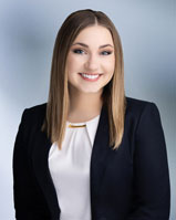 Attractive blond women, Lindsey Mead, smiles at camera wearing dark blue sport coat and white blouse in front of blurred blue gray background