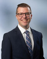 Man with glasses, Taylor Gast, Smiles at Camera Wearing a dark blue suit in front of blurred blue gray background 