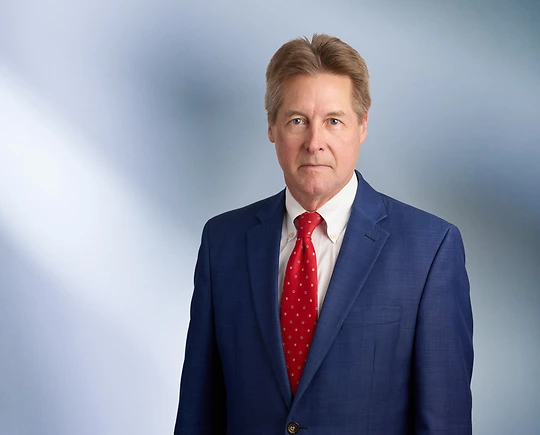 A man, James Doezema, in a dark suit, light shirt, and striped tie is smiling at the camera, standing against a blurred blue and white background.