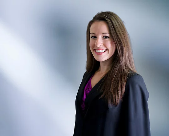 A woman, Julie Hamlet, in a black blazer and purple top is smiling at the camera, standing against a blurred blue and white background.