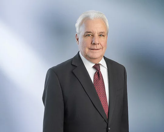 man with white hair wearing a dark suit, white shirt, and maroon patterned tie, standing against a softly blurred blue and white background