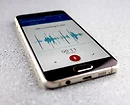 A smartphone lying on a gray surface displaying a paused voice recording app, with visible audio waveforms and a red record button.