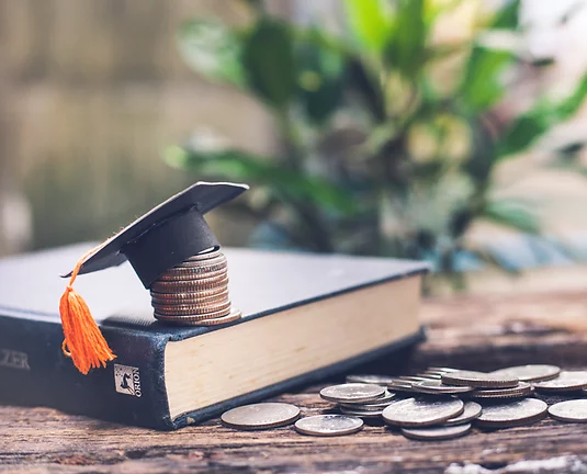 Mini graduation cap on stack of coins on top of book which is on desk near second pile of coins
