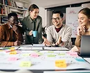 Four diverse coworkers collaborating at a table covered in colorful sticky notes and planning charts. One person is talking and gesturing, while others listen and smile in a creative office setting.