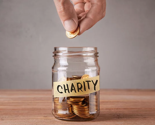 A hand placing a gold coin into a glass jar labeled "CHARITY," which is partially filled with similar coins, symbolizing a donation or charitable contribution.