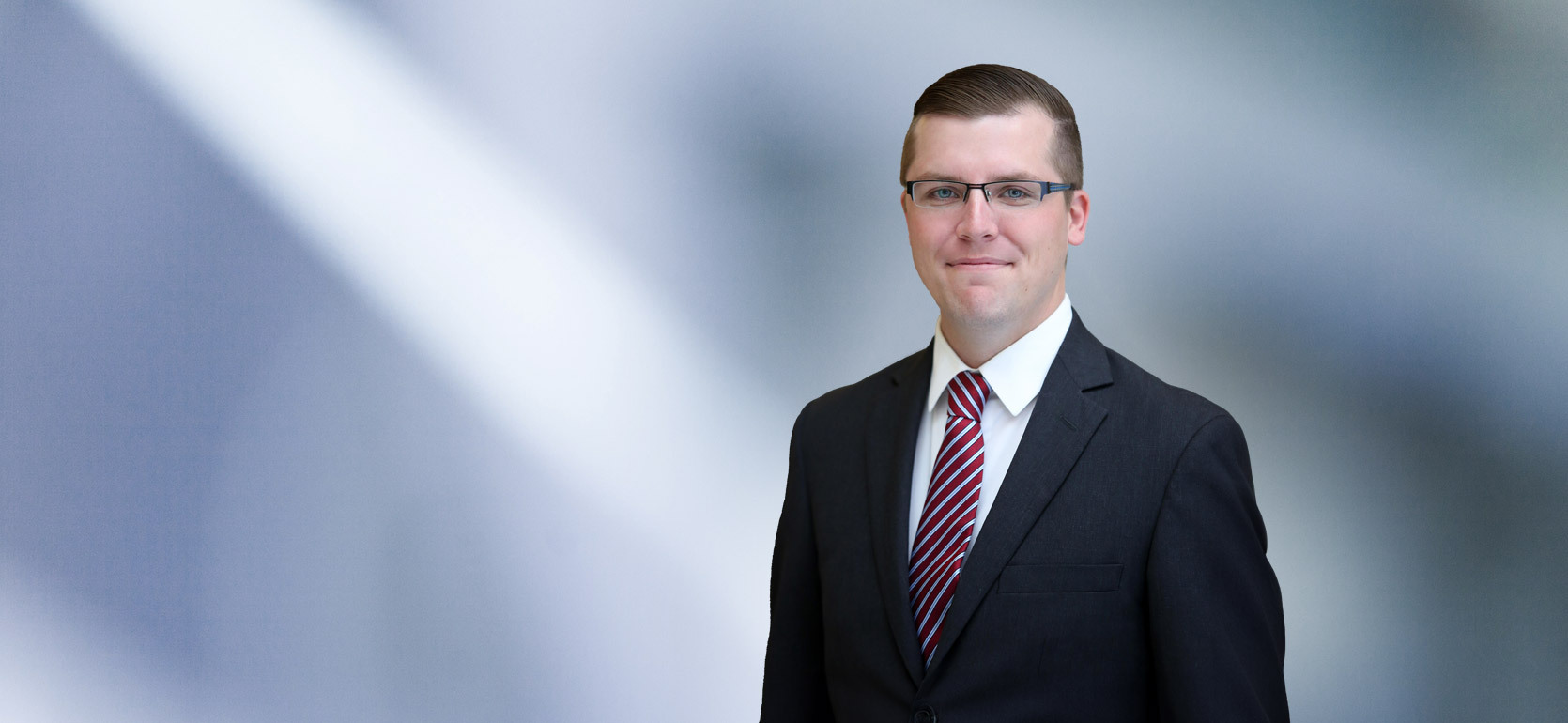 man, Taylor Gast, in a dark suit smiling at the camera, standing against a blurred blue and white background.