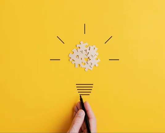 Paper puzzle pieces with hand drawn lines around to look like a lightbulb on a mustard yellow background.