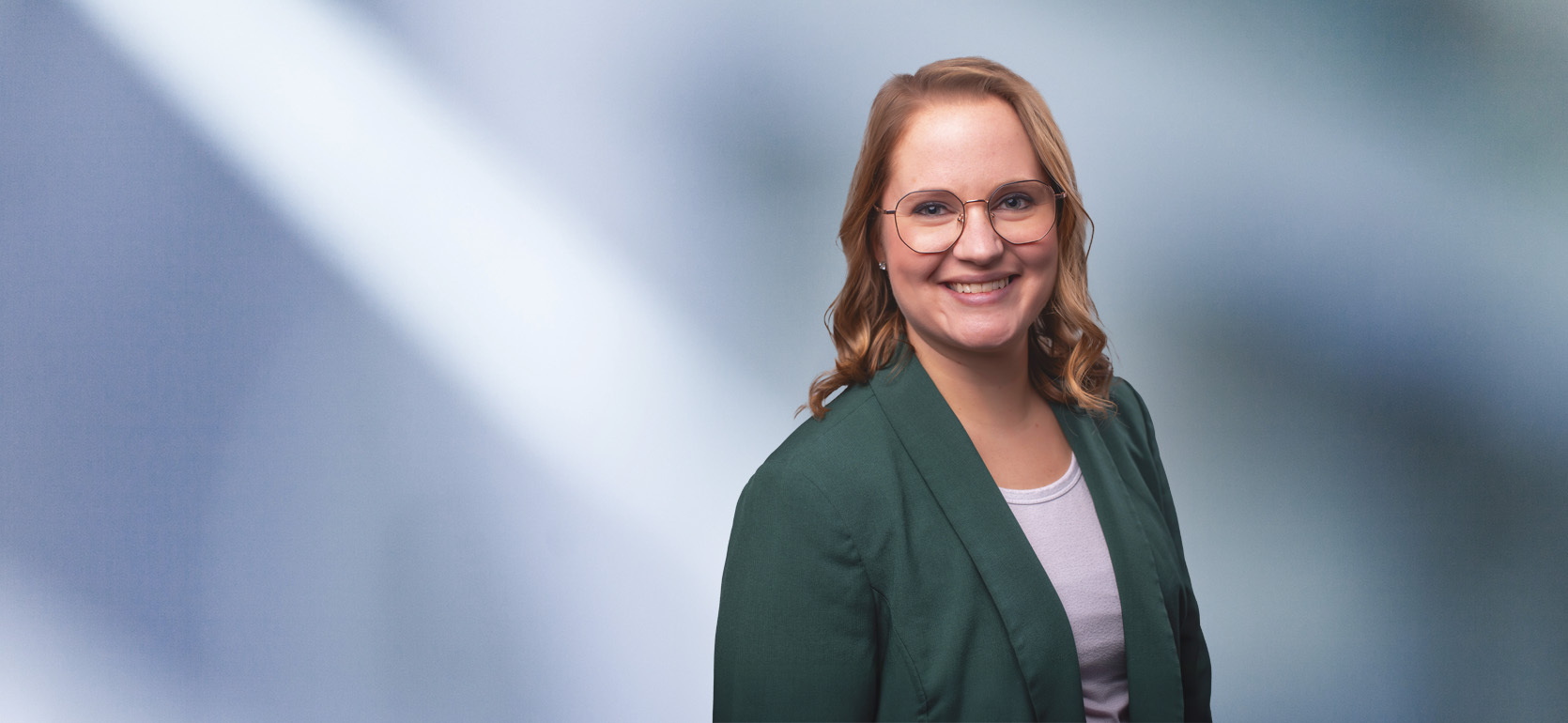 “A woman, Megan Cochrane, with shoulder-length, straight blonde hair, wearing a blue blazer and white top, is smiling at the camera, standing against a blurred blue and white background.”