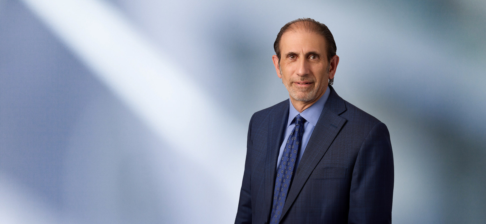 A man, Jonathan David, in a dark blue suit, light blue shirt, and patterned tie is looking at the camera, standing against a blurred blue and white background.
