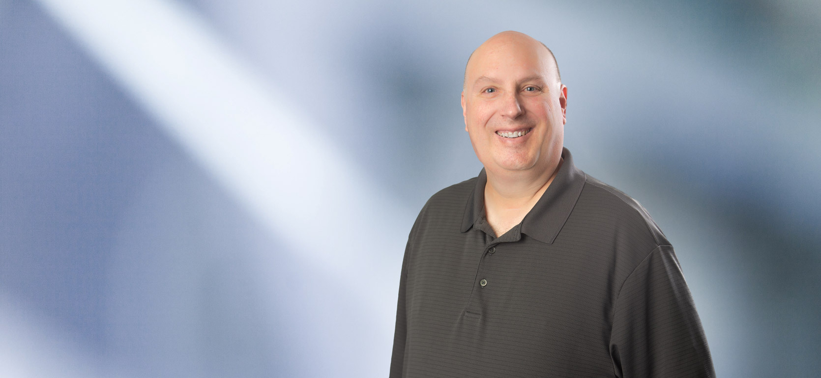 Smiling bald man, John Bonandrini, wearing a dark grey polo shirt, standing in front of a soft blue gradient background.