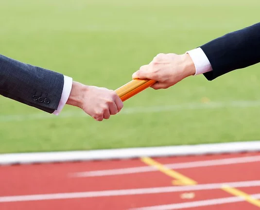 A hand handing a track baton to another hand with a track field in the background.