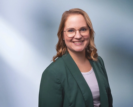 “A woman, Megan Cochrane, with shoulder-length, straight blonde hair, wearing a blue blazer and white top, is smiling at the camera, standing against a blurred blue and white background.”