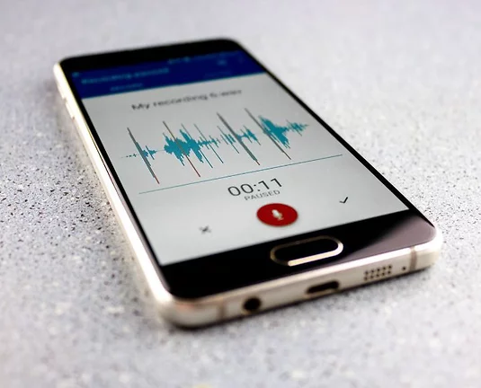 A smartphone lying on a gray surface displaying a paused voice recording app, with visible audio waveforms and a red record button.