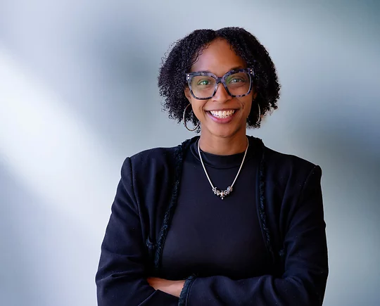 African American woman, Kanielle Pounds, with large black rimmed glasses smiles at camera in front of blue gray background wearing a black sweater and black shirt