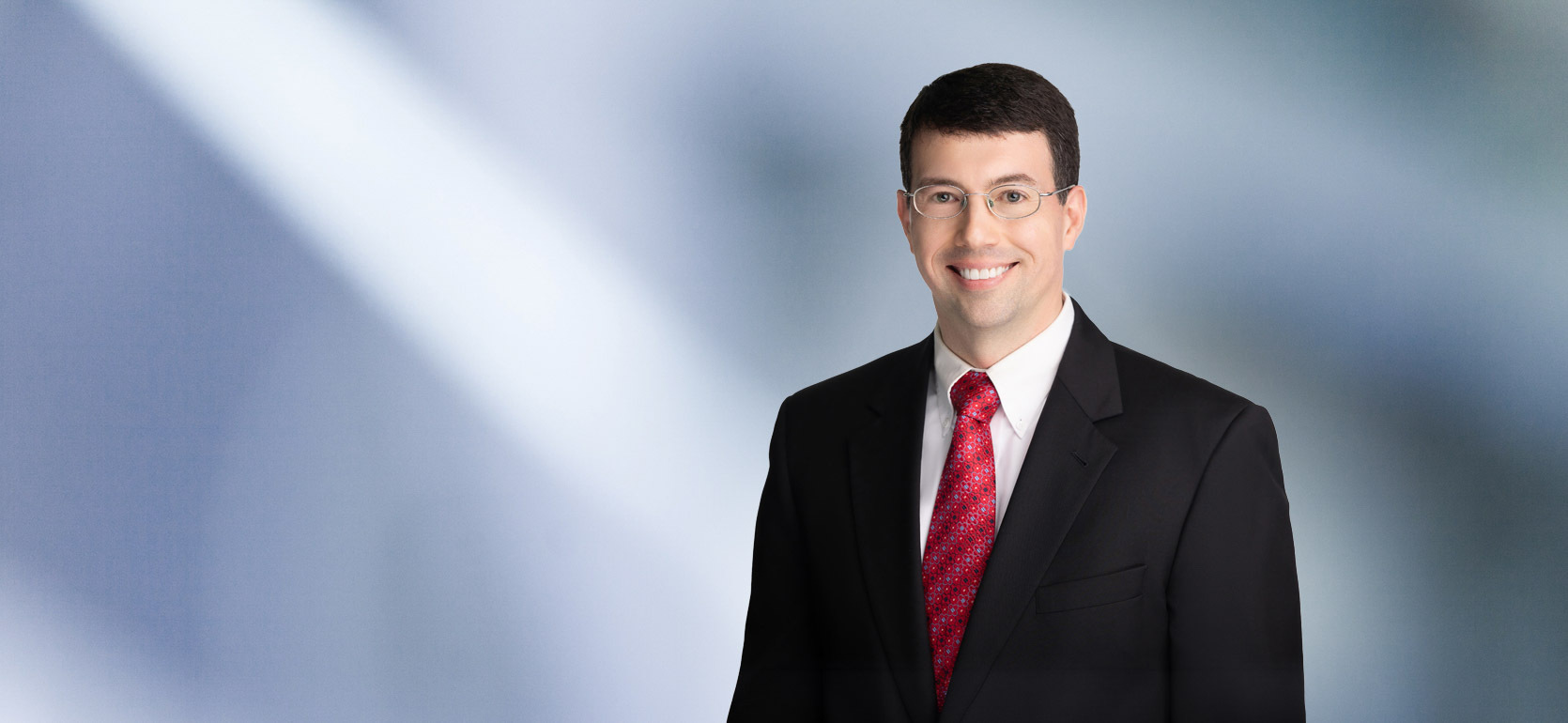 A man, James Anderton, in a black suit, white shirt, and red patterned tie is smiling at the camera, standing against a blurred blue and white background.