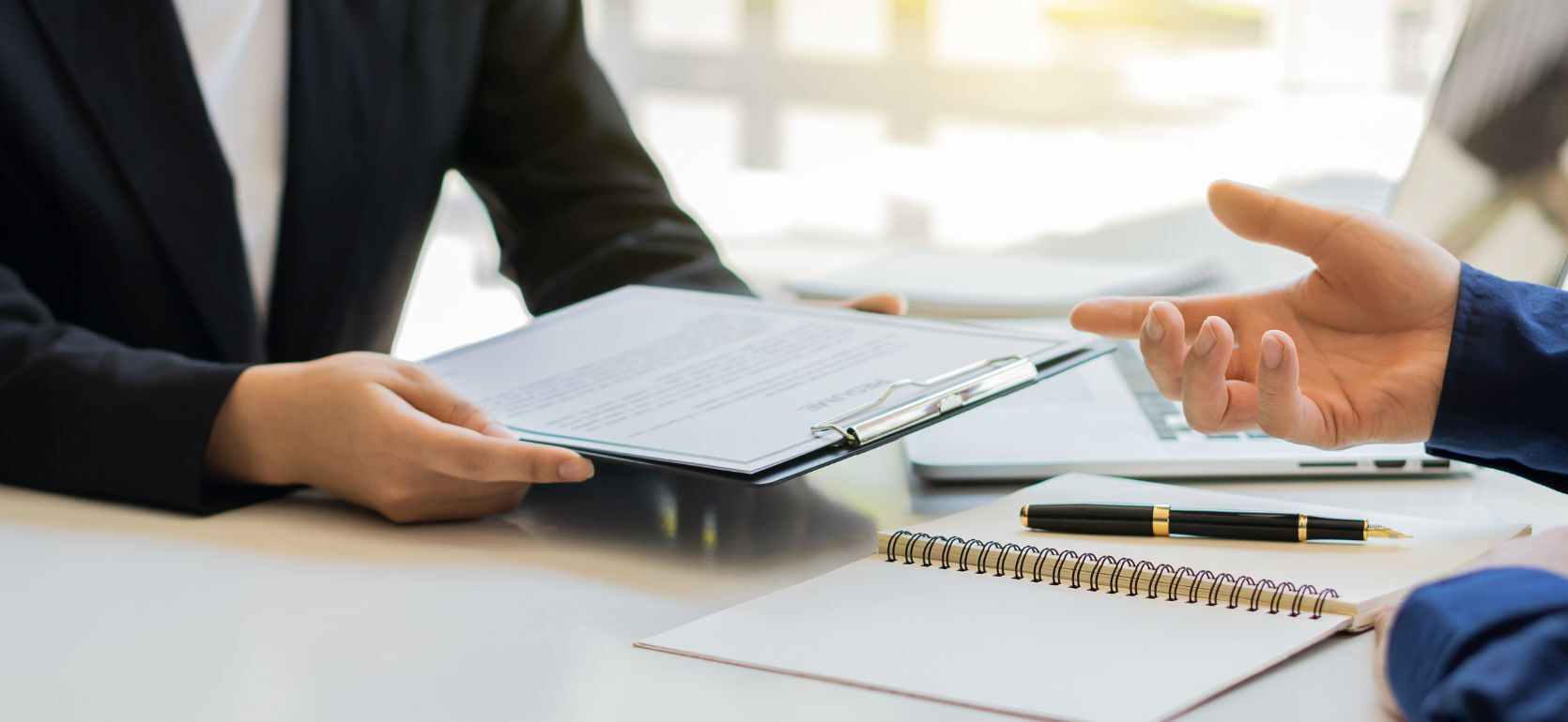 Two people discussing while one person has a clipboard and papers and a pen on a desk.