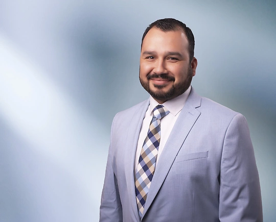 A man, Juan Negrete, in a light blue suit, white shirt, and patterned tie is smiling at the camera, standing against a blurred blue and white background.
