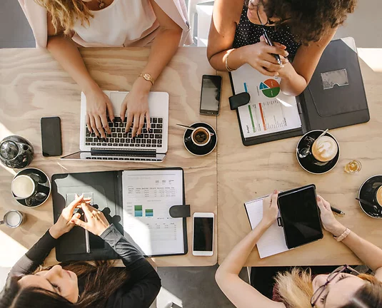 Four businesswomen sit around table with cups of coffee and laptops and documents