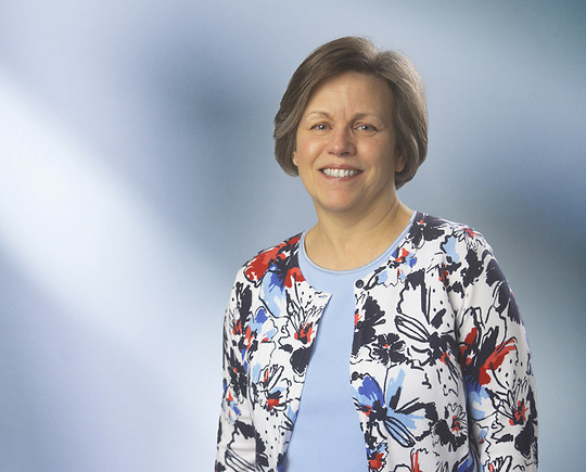 A woman, Lori Rosier, with straight, blonde hair, wearing a black blazer and white top, is smiling at the camera, standing against a blurred blue and white background.”