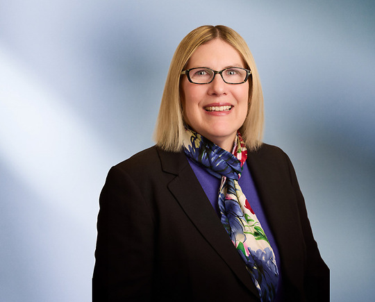 A woman, Jennifer Vanregemorter, in a black blazer, purple top, and multicolored floral scarf is smiling at the camera, standing against a blurred blue and white background.