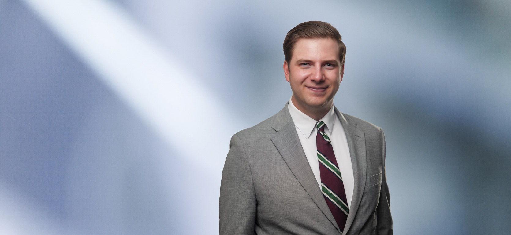 “A man, Matthew Murray, with short, light brown hair, wearing a grey blazer and white shirt, is smiling at the camera, standing against a blurred blue and white background.”