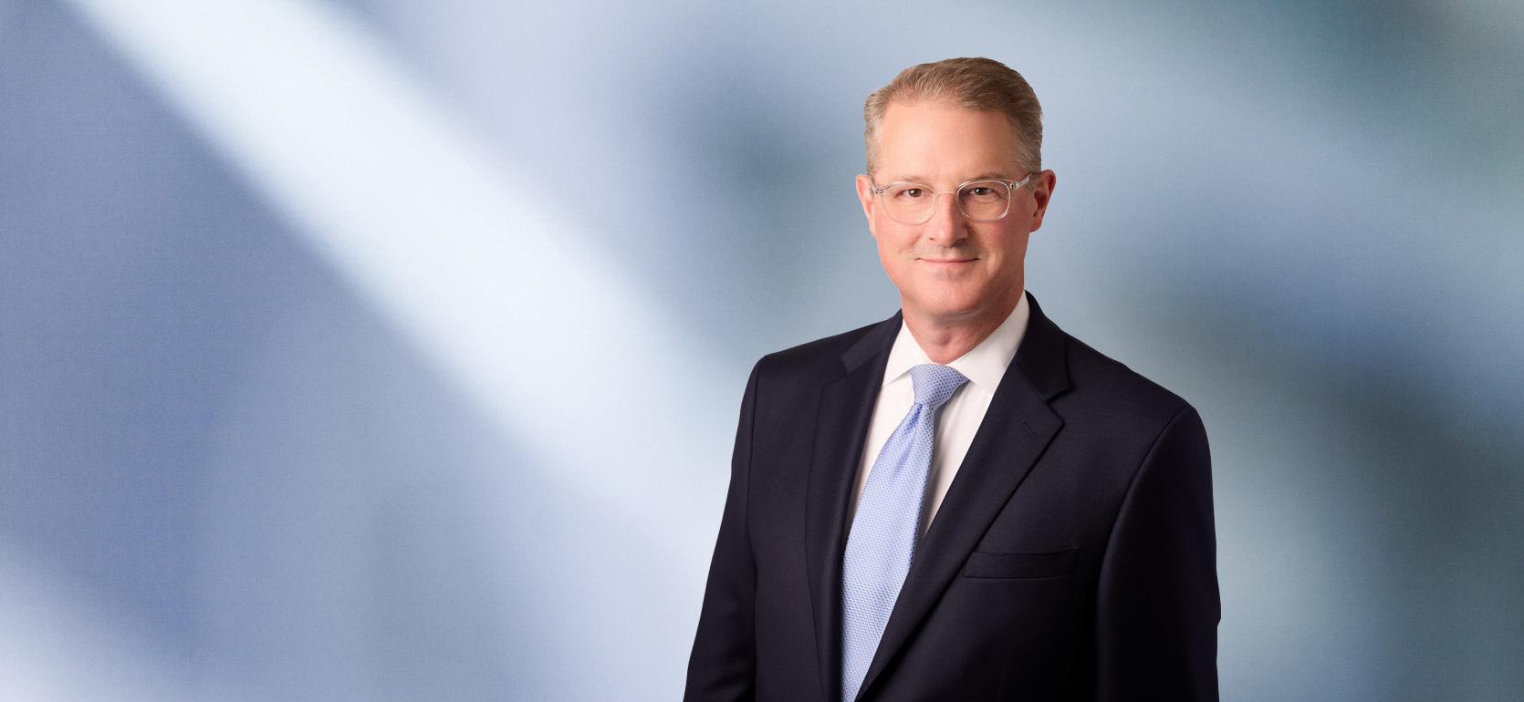 Portrait of a man, Thomas TerMaat, with short white hair in a dark blazer, with a blurred blue and white background.