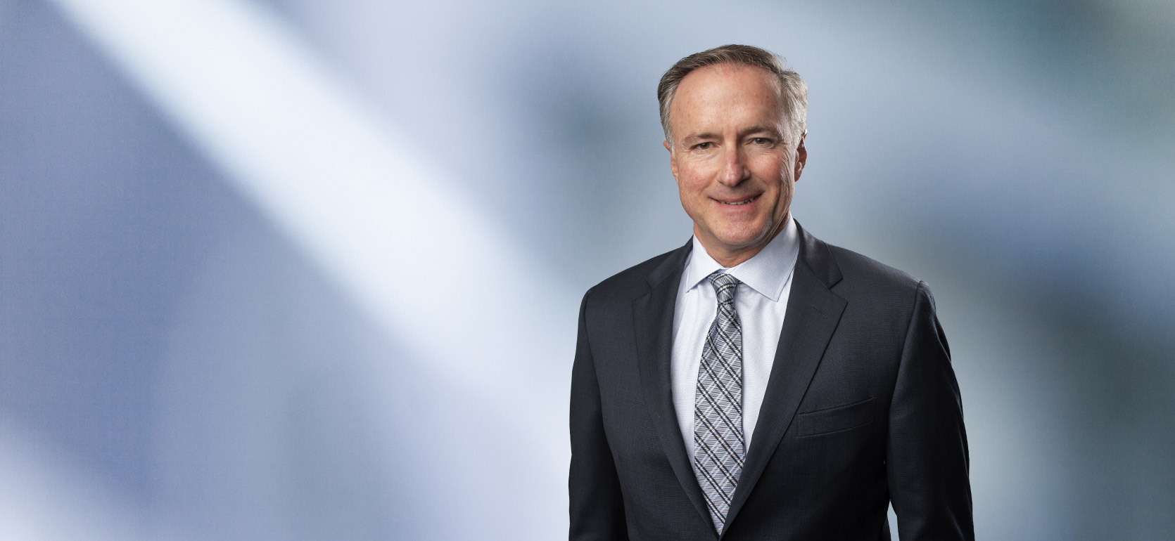 A man, Paul Millenbach, in a dark suit, light shirt, and patterned tie is smiling at the camera, standing against a blurred blue and white background.