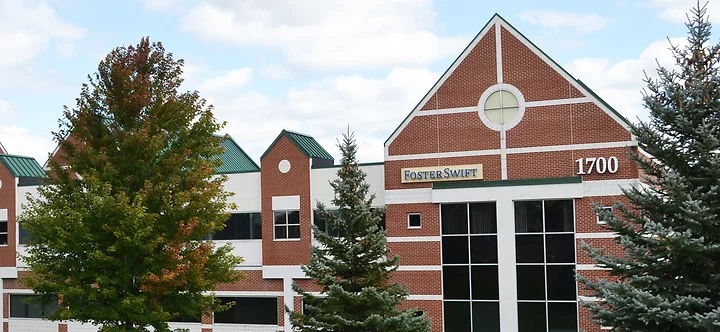 Exterior of the Foster Swift Grand Rapids office building. The structure features red brick with white trim, green peaked roofs, and large windows. A sign reading "Foster Swift" is mounted near the building's peak, alongside the number 1700.