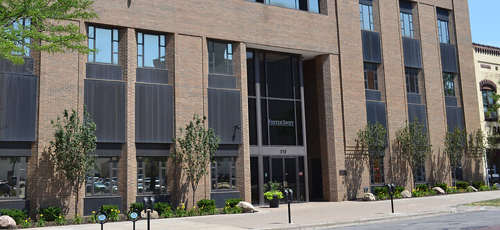 Exterior view of the Foster Swift Collins & Smith PC Lansing office building. The brick facade features tall vertical windows with dark paneling, a glass entrance with the firm's name above the door, and a landscaped sidewalk with young trees and flower b
