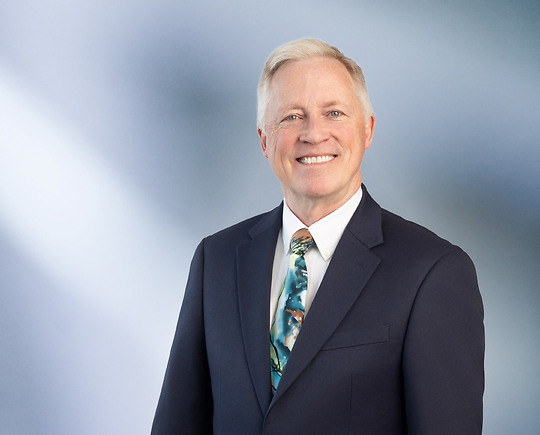 Professional man, Michael Liddane, with light hair wearing a dark suit, white shirt, and colorful patterned tie, standing against a softly blurred blue and white background