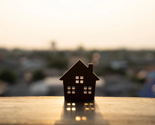Silhouette of a small wooden house model on a ledge at sunset, with a blurred cityscape in the background