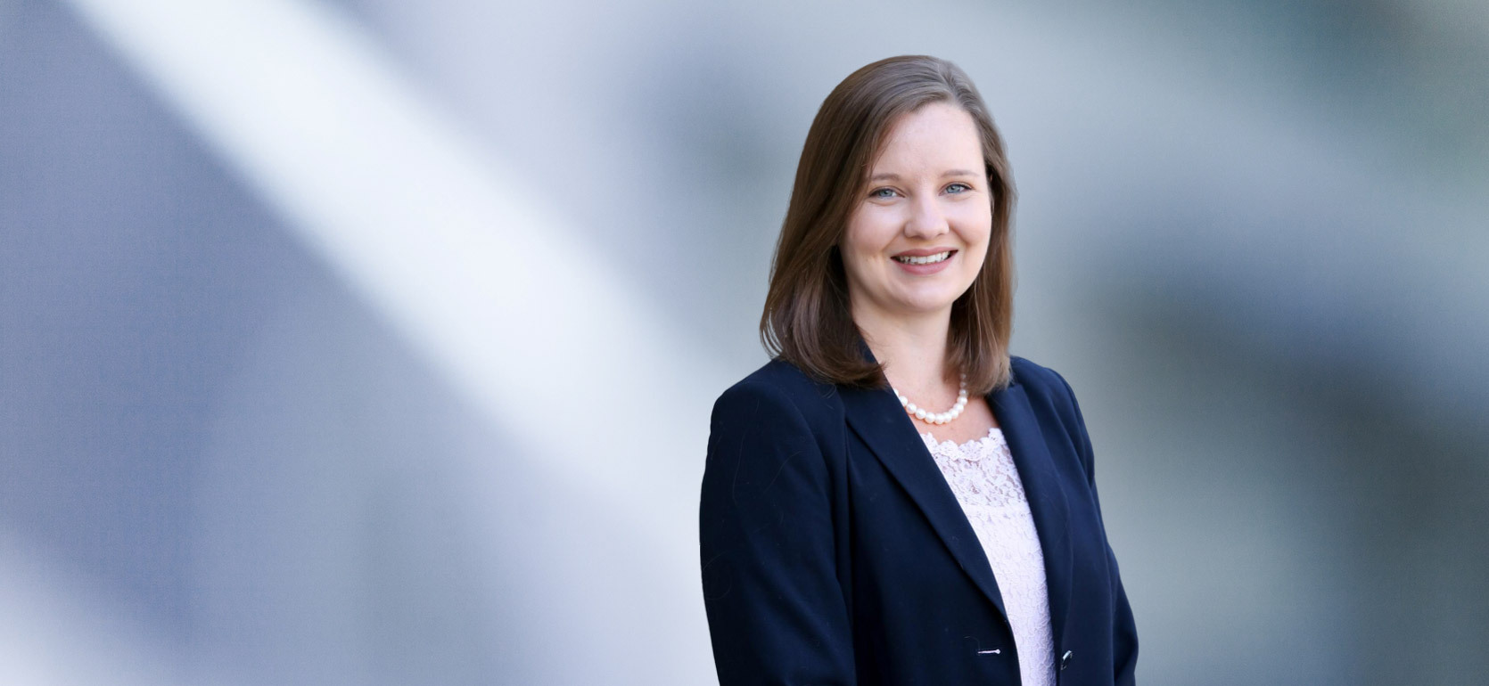 Smiling woman, Allison Collins, with shoulder-length light brown hair, wearing a dark blazer, white lace blouse, and pearl necklace, standing in front of a soft blue gradient background