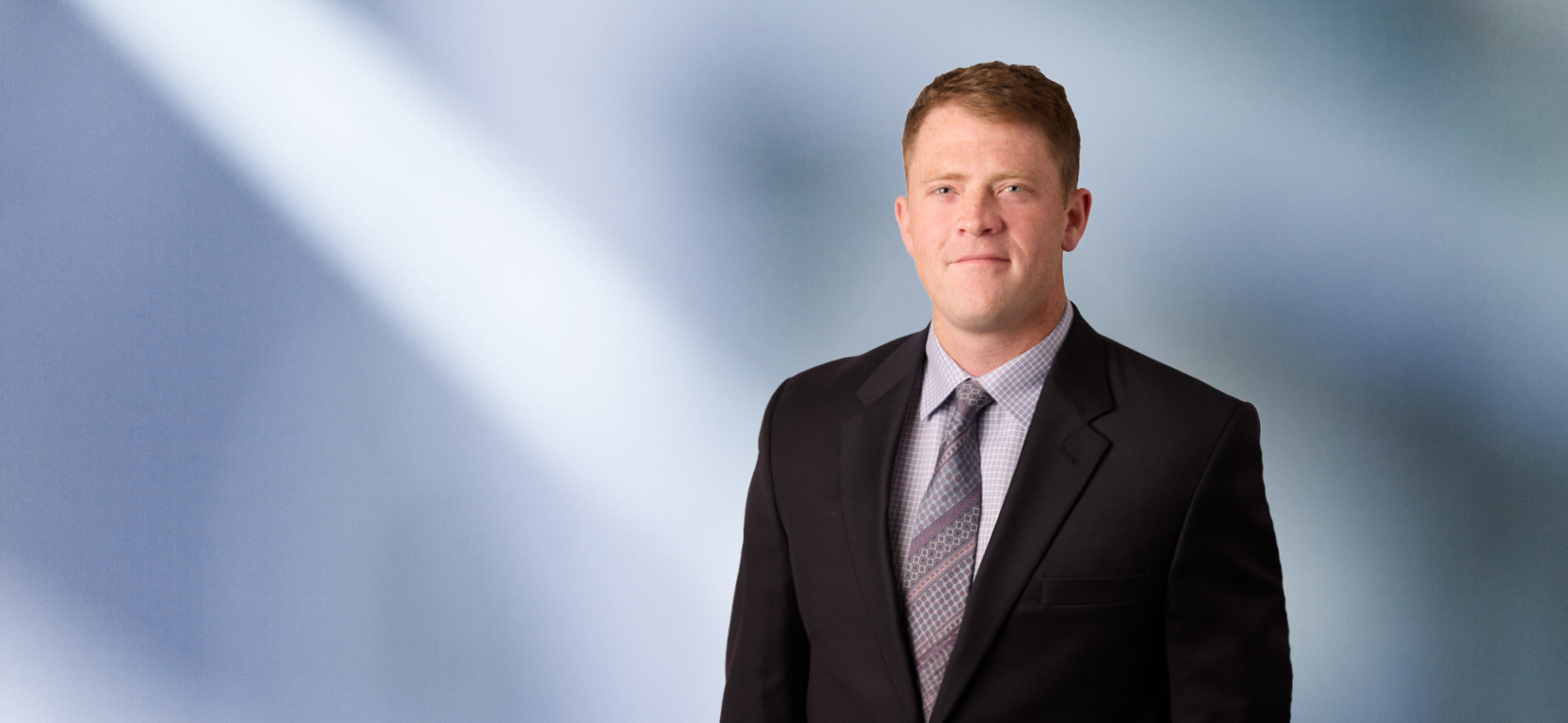 Man, Nicholas Stock, with short dark brown hair wearing a blue blazer, smiling against a blurred white and blue background.