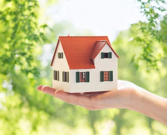 Model of a white cottage with an orange roof and green shutters in a hand with trees in the background.