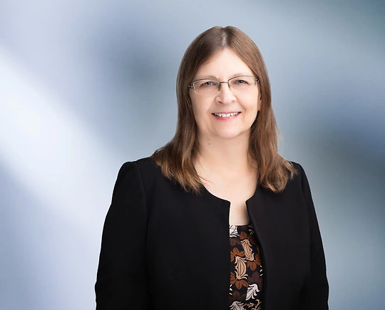 A woman, Beverly Swan, in a black blazer and patterned top is smiling at the camera, standing against a blurred blue and white background.