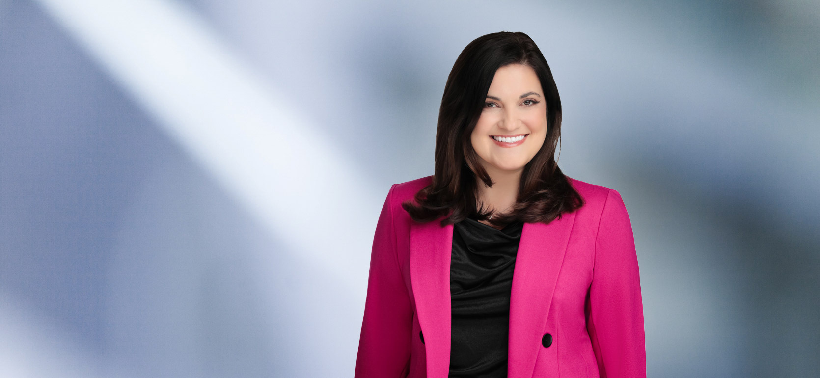 Woman, Stefania Gismondi, wearing a pink blazer, standing against a blurred blue and white background.