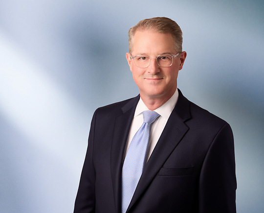 Portrait of a man, Thomas TerMaat, with short white hair in a dark blazer, with a blurred blue and white background.