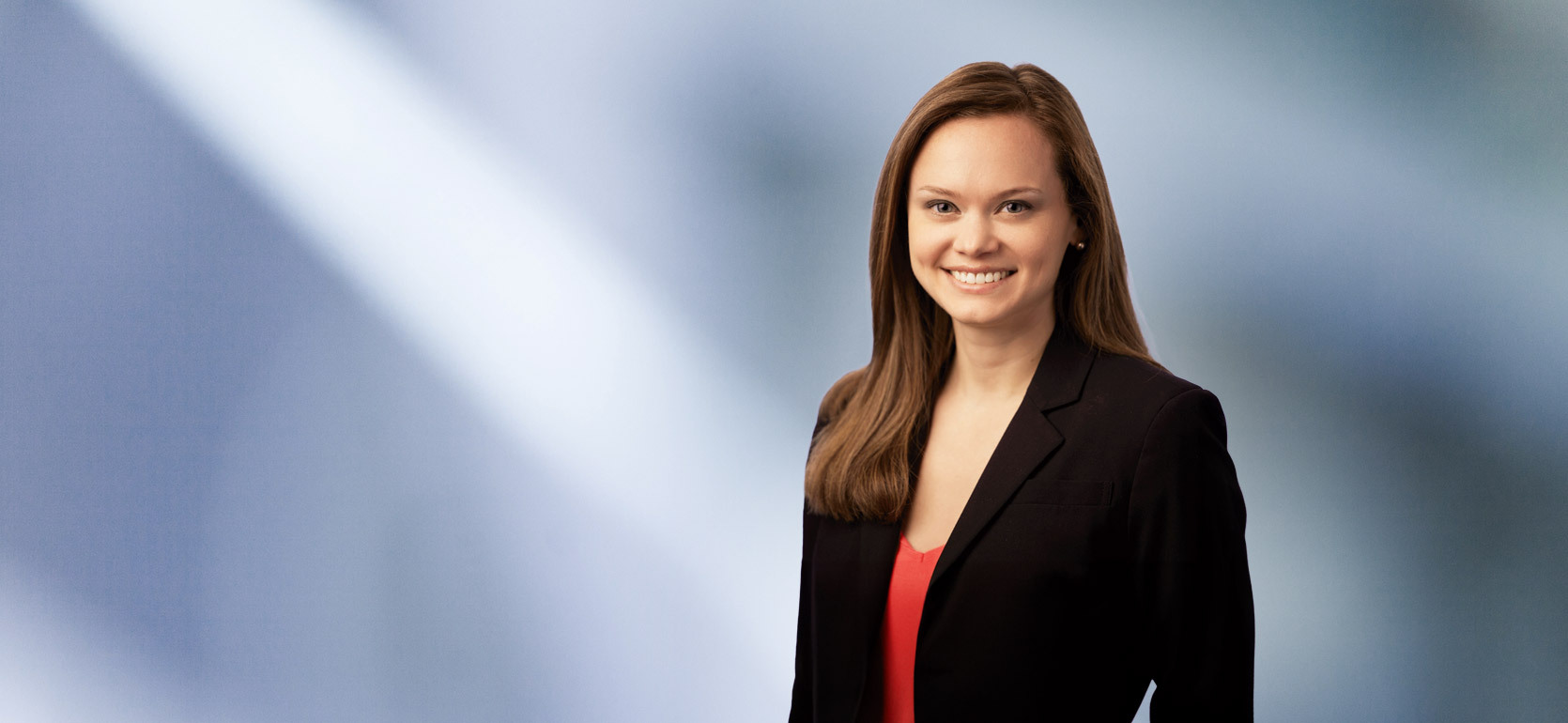 A woman, Leslie Abdoo, in a black blazer and red top is smiling at the camera, standing against a blurred blue and white background.