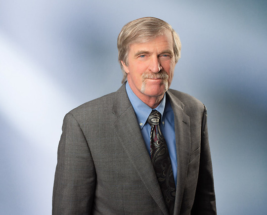 Portrait of a man, Steven Tjapkes, with short light gray hair in a dark blazer, with a blurred blue and white background.