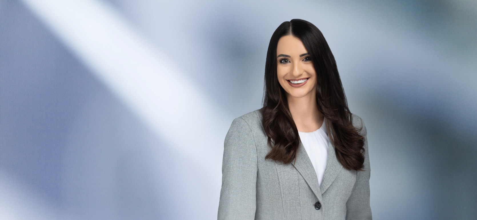 “A woman, Melanie Assad, with long, dark brown hair, wearing a grey blazer and white top, is smiling at the camera, standing against a blurred blue and white background.”