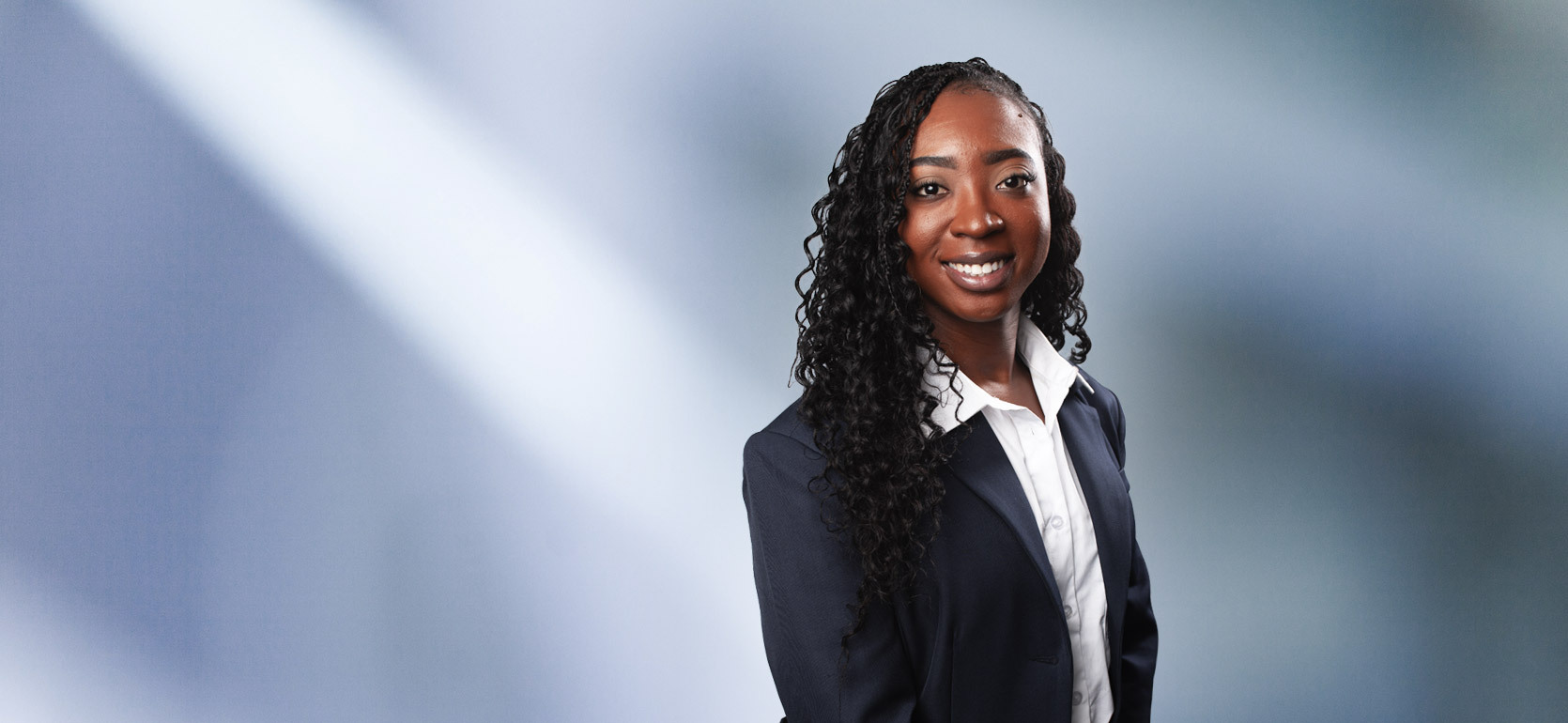 A woman, Kendra Dixie, in a dark navy blazer over a white button-up shirt is smiling at the camera, standing against a blurred blue and white background.