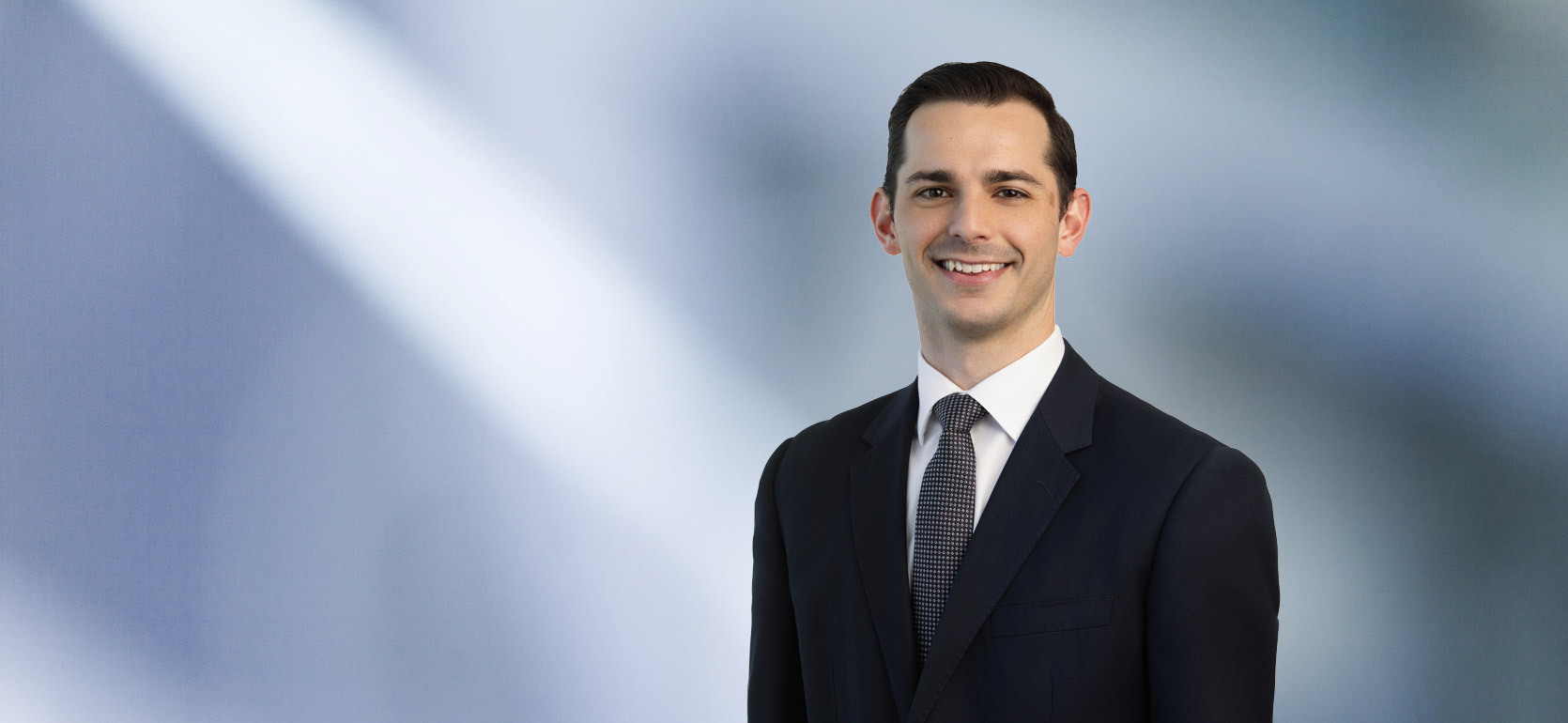 man, Lino Taormina, wearing a dark suit, white shirt, and patterned tie, standing against a softly blurred blue and white background, smiling confidently at the camera.